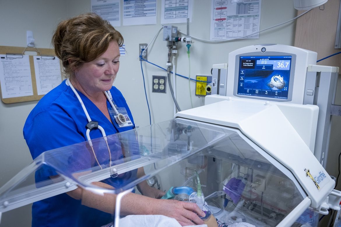 Julie Vanyolai is pictured caring for a young patient in an incubator/isolette. She has strawberry blonde/red hair, which is tied up in a bun, and blue eyes. She’s wearing blue scrubs and has a stethoscope around her neck.