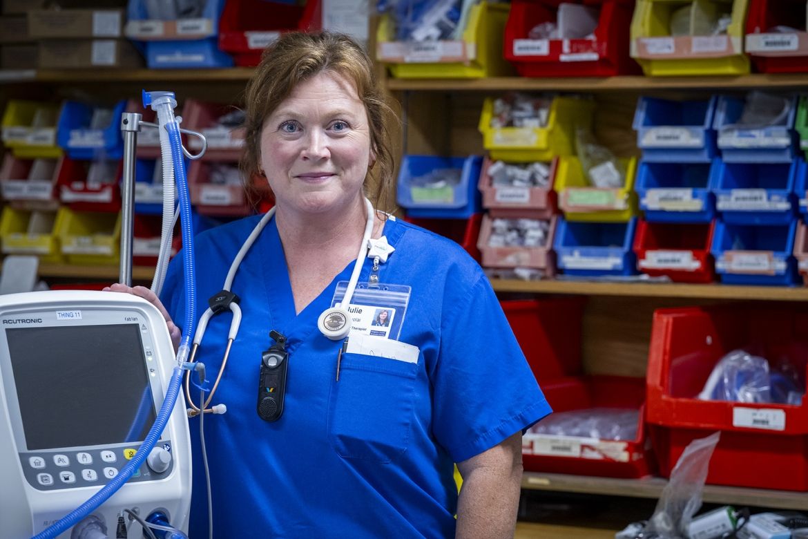 Julie Vanyolai is pictured standing next to a piece of respiratory therapy equipment, and is looking directly into the camera. She has strawberry blonde/red hair, which is tied up in a bun, and blue eyes. She’s wearing blue scrubs and has a stethoscope around her neck.