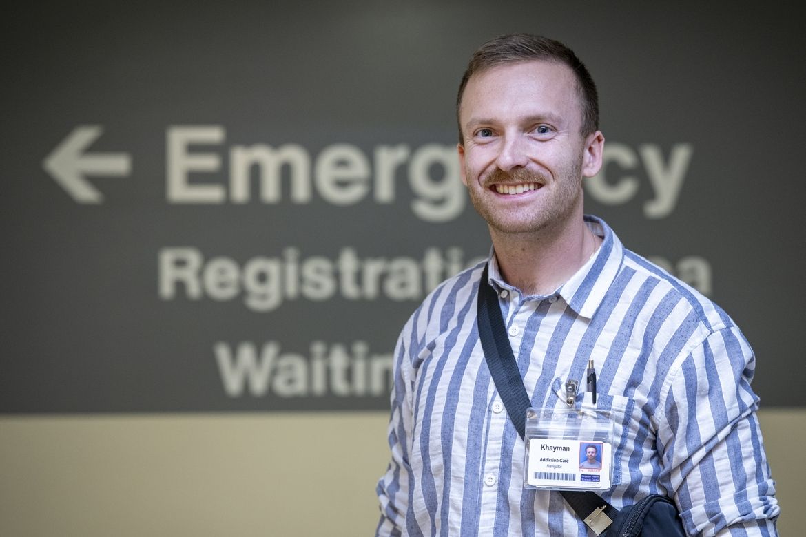 Khayman Wood is wearing a striped shirt and his KHSC ID badge, while standing smiling directly to the camera in a hallway outside the Emergency Department. The sign in the background has the words "emergency," "registration area," and "waiting" partially visible, with an arrow pointing to the left.