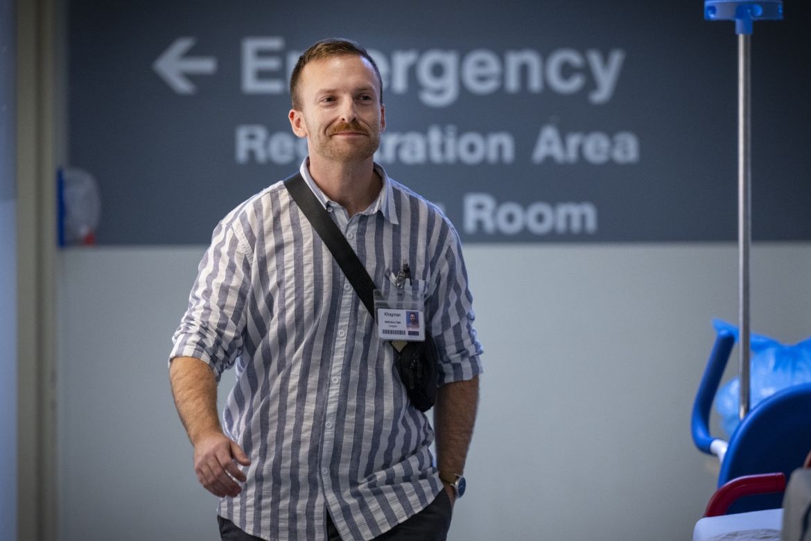 Khayman Wood is walking down a hallway at the Kingston General Hospital site. Behind him, a sign reads "emergency registration area room" with an arrow pointing to the left. A blue medical chair and equipment are partially visible on the right side of the image.
