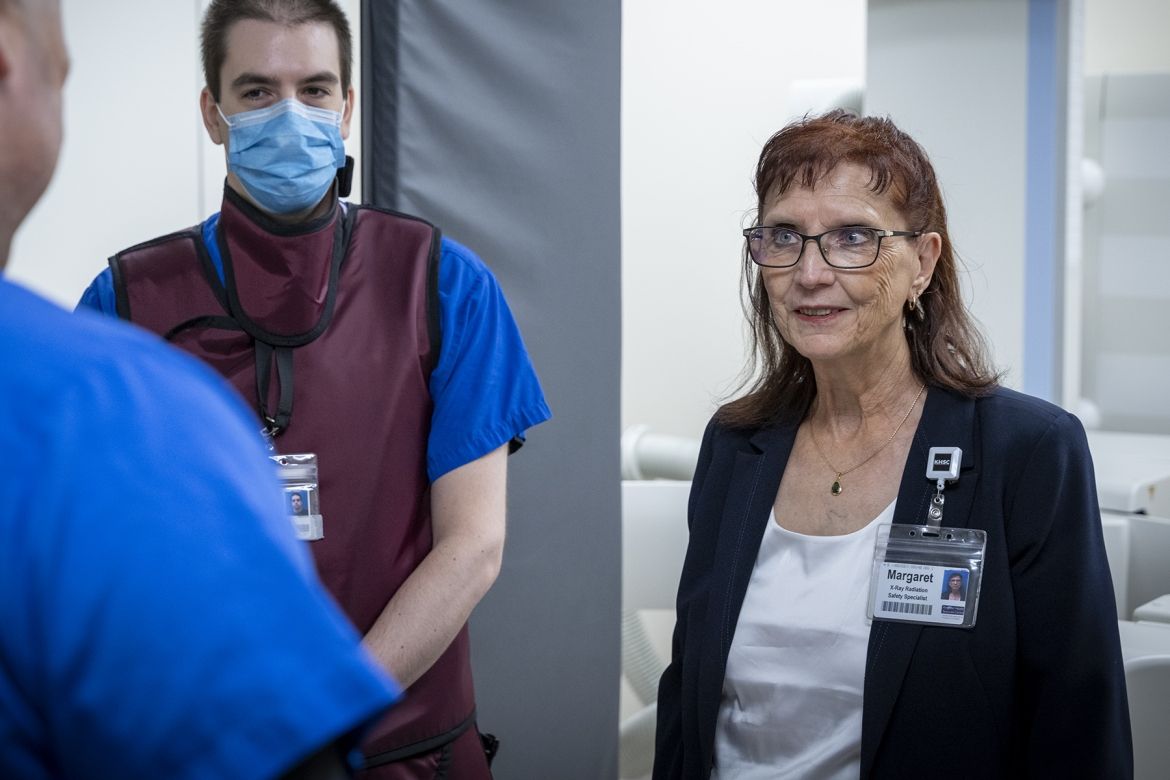 Margaret Bollen is pictured standing in a suite at Kingston General Hospital. She has auburn hair, blue eye hair and wears glasses. She’s wearing a dark navy suit and white top. She’s standing next to a medical radiation technologist.