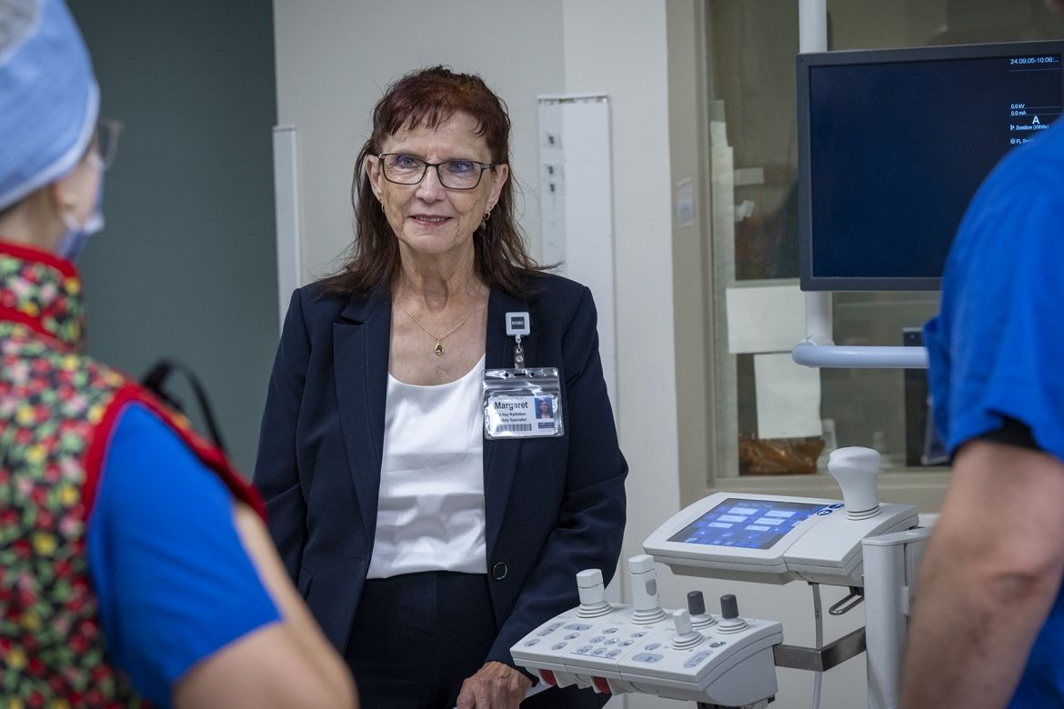 Margaret Bollen is pictured standing in a suite at Kingston General Hospital, next to a piece of equipment. She has auburn hair, blue eye hair and wears glasses. She’s wearing a dark navy suit and white top. She’s chatting with a medical radiation technologist.