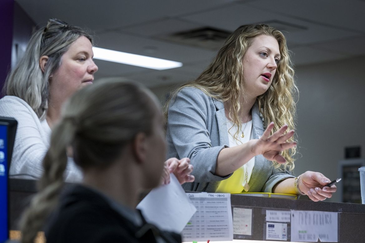 Orlana Bourgoin is standing and leaning up against a care desk in an inpatient unit at the Kingston General Hospital site. She’s talking to two other staff members featured in the image. She has long, wavy, blonde hair, blue eyes and is wearing a grey blazer with a white blouse underneath.