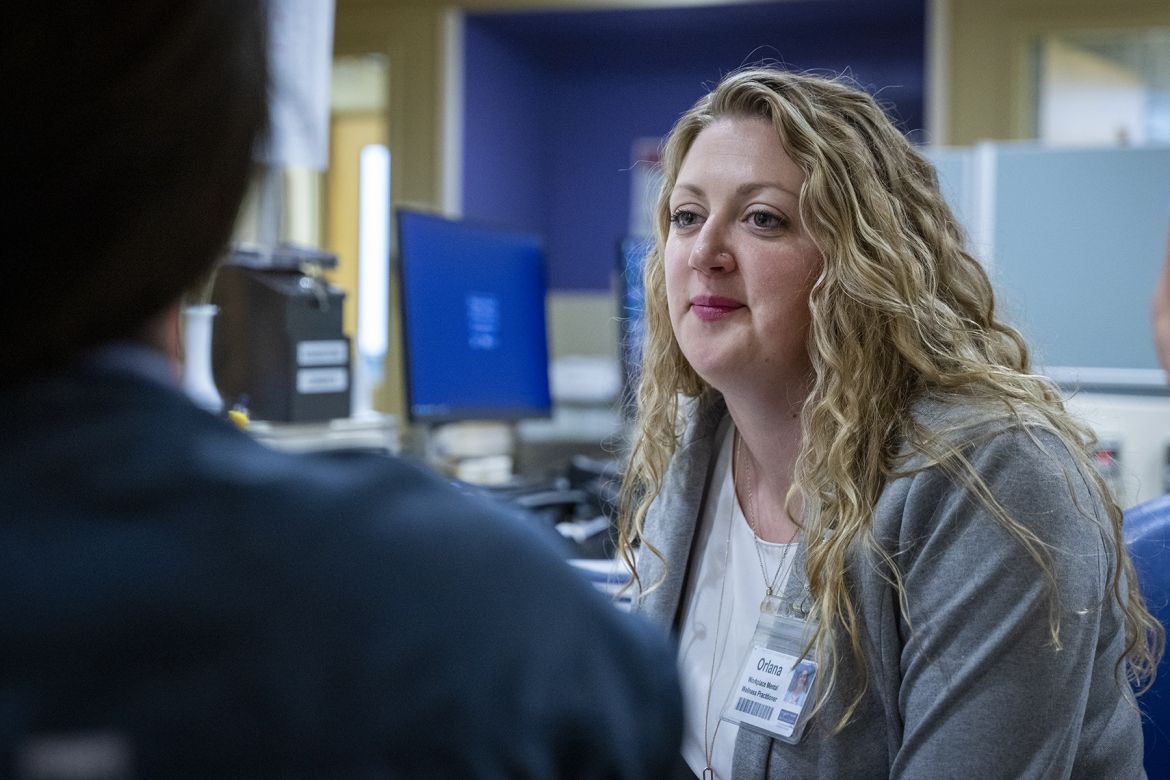 Orlana Bourgoin is sitting on a chair at a care desk in an inpatient unit at the Kingston General Hospital site, looking at a colleague. She has long, wavy, blonde hair, blue eyes and is wearing a grey blazer with a white blouse underneath.
