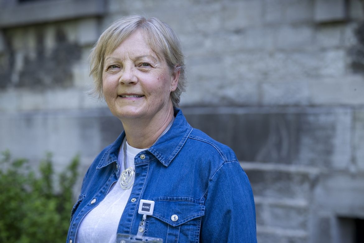 Patti Cox is pictured smiling directly at the camera while standing outside at Kingston General Hospital. She has blue eyes and light blonde hair styled in a pixie cut. She’s wearing a blue denim blouse on top of a white t-shirt.