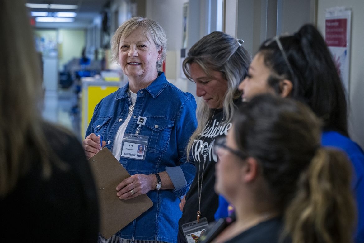 Patti Cox is pictured taking part of team huddle on a patient care unit. She’s smiling as a member of the care team speaks to the group. Cox has blue eyes and light blonde hair styled in a pixie cut. She’s wearing a blue denim blouse on top of a white t-shirt, and is holding a clipboard and pen.