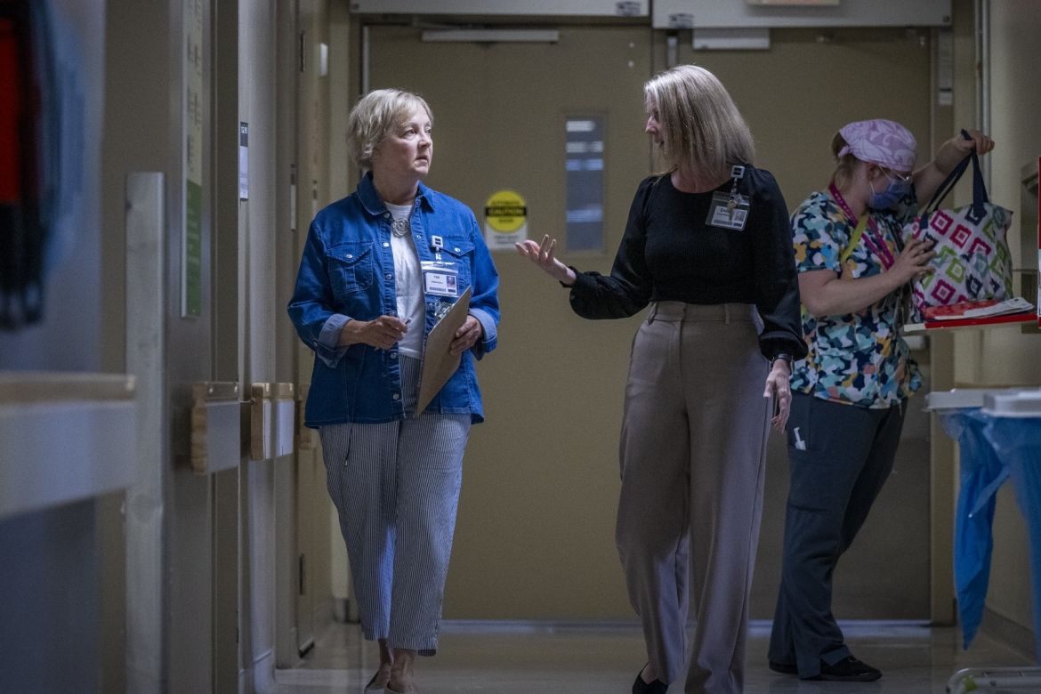 Patti Cox is pictured walking and talking with a member of patient care team down a hallway on an in-patient unit. Cox has blue eyes and light blonde hair styled in a pixie cut. She’s wearing a blue denim blouse on top of a white t-shirt, and is holding a clipboard and pen. The care team member has should length blonde hair, and is wearing a black blouse with light brown dress pants.
