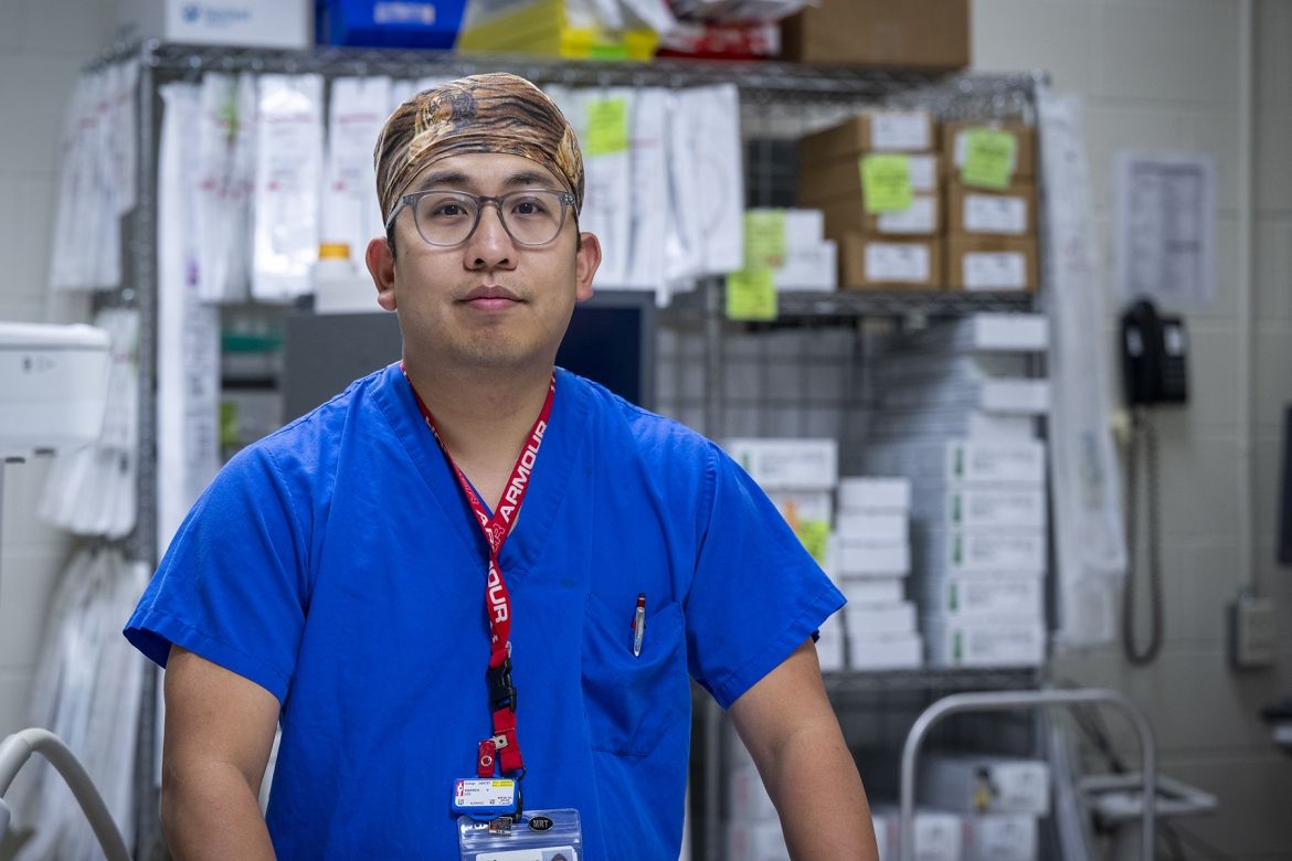 Warren Lee is pictured standing in room filled with medical equipment at the Kingston General Hospital site. He’s facing the camera directly and wearing bright, blue scrubs, a tiger print scrub cap, and has grey, translucent glasses.