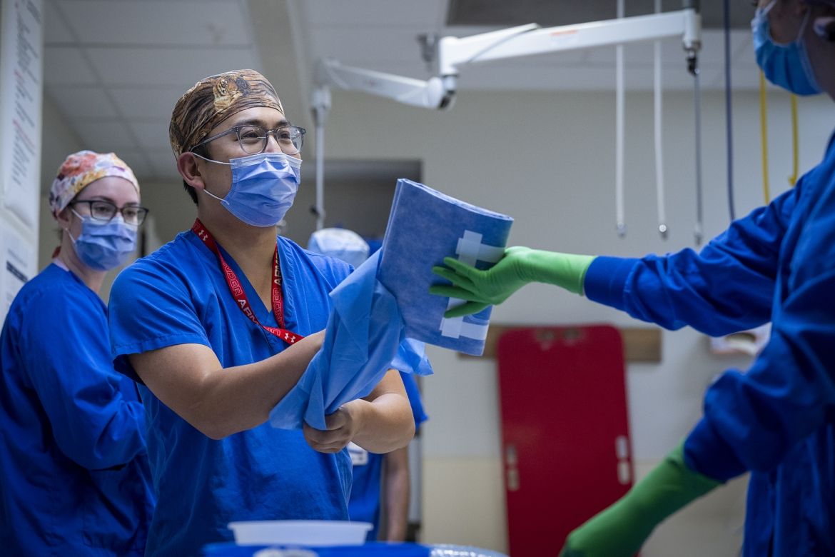 Warren Lee is pictured in a room with two other colleagues, who are getting ready for a procedure. Lee is wearing bright, blue scrubs, tiger print scrub cap, a mask over his nose and mouth, and has grey, translucent glasses. He’s facing a colleague who is handing him some blue clothes.