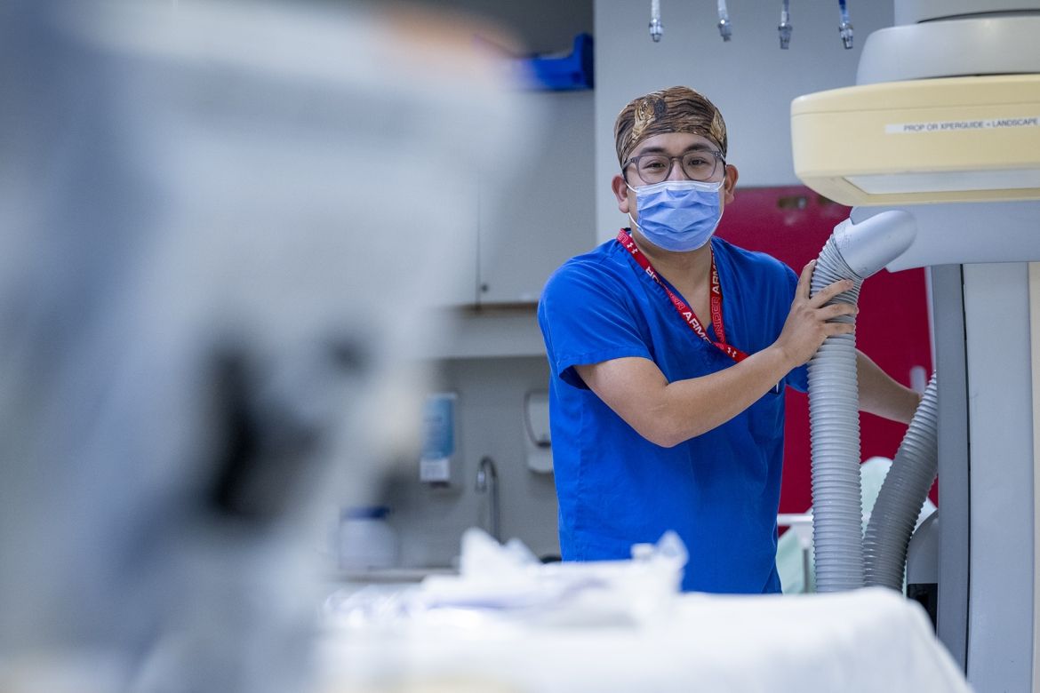 Warren Lee is pictured in the background of an image next to a piece of equipment. He’s holding the equipment while looking directly at the camera. Lee is wearing bright, blue scrubs, tiger print scrub cap, a mask over his nose and mouth, and has grey, translucent glasses.