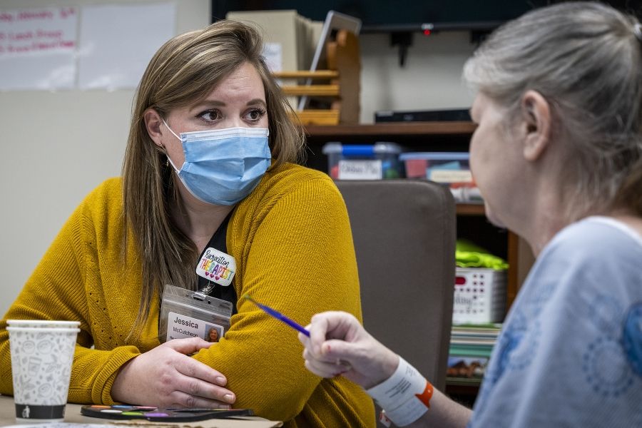 Jessica McCutcheon is seated next to an elderly female patient, watching her do arts and crafts. McCutcheon has dark blonde hair and brown eyes. She’s wearing a black top, mustard cardigan and a mask over her mouth and nose.