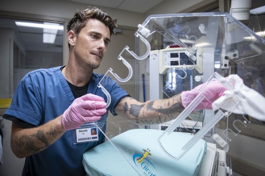 Feature image: Dennis Clark is pictured in the NICU at the KGH site, cleaning equipment. He has short brown hair, blue eyes and has tattoos on both arms. He’s wearing blue scrubs.