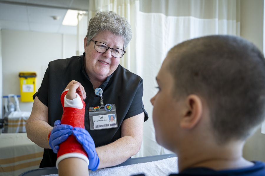 Teri Timpany is putting a red cast on a young patient. She’s smiling and looking at him, while she puts a cast on his arm. Teri has short, curly, silver hair and wears glasses. She’s wearing black scrubs.