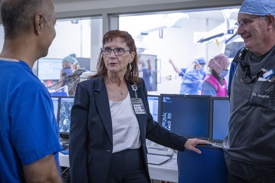 Margaret Bollen is pictured standing in a suite at Kingston General Hospital. She has auburn hair, blue eye hair and wears glasses. She’s wearing a dark navy suit and white top. She’s chatting with physician and a medical radiation technologist.