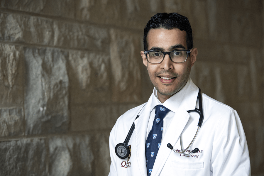 Dr. Aws Almufleh is pictured standing in a hallway at the Hotel Dieu Hospital site. He has short, dark hair, dark brown eyes, has glasses, and is pictured wearing a white laboratory coat.