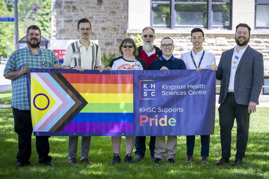 Seven members of the Pride Staff Community Group are standing outside Kingston General Hospital, holding a large Progress Pride banner that says Kingston Health Sciences Centre supports Pride.