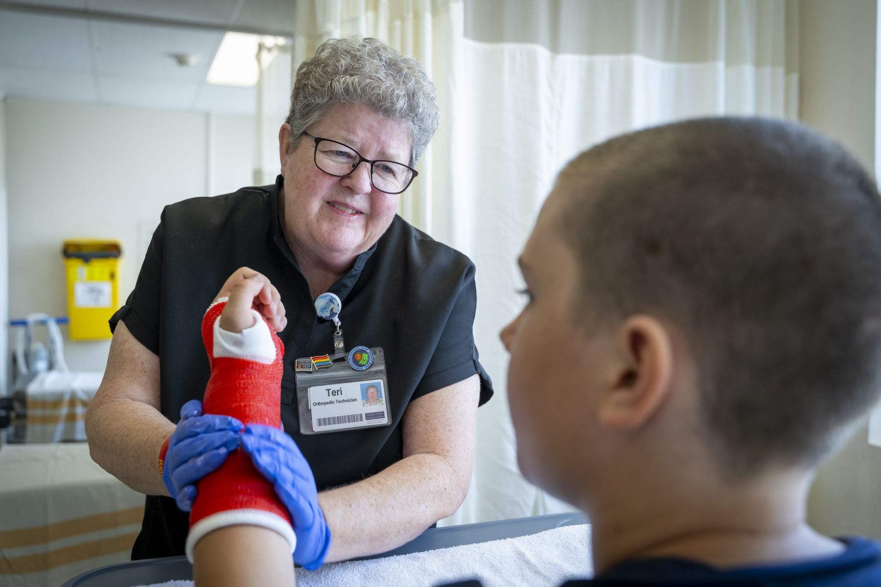 Teri Timpany is putting a red cast on a young patient. She’s smiling and looking at him, while she puts a cast on his arm. Teri has short, curly, silver hair and wears glasses. She’s wearing black scrubs.
