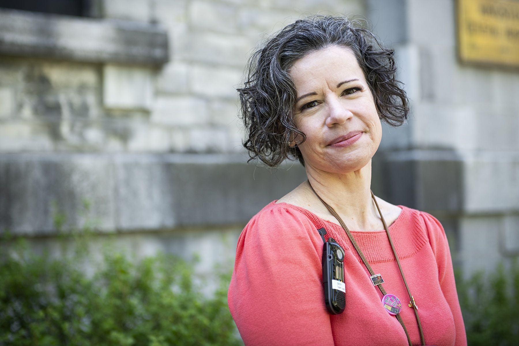 Jane Lewis has short, dark and wavy hair, and dark brown eyes. She’s wearing a bright coral blouse and is pictured standing outside Kingston General Hospital.  